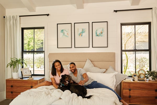 Interior of white, modern bedroom with a couple and dog sitting on bed in front of black framed Andersen Windows