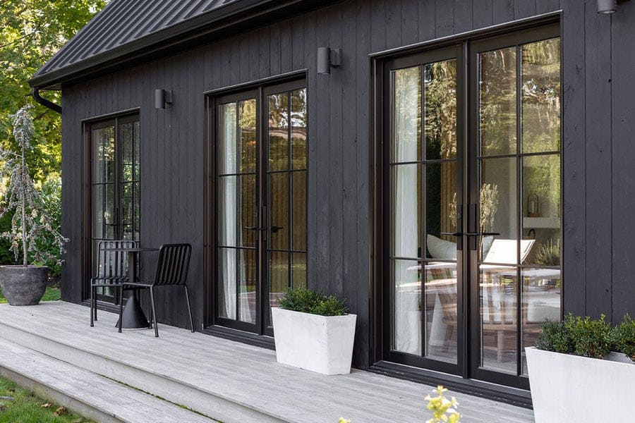 An exterior shot of a black barn-inspired addition featuring three sets of black E-Series Hinged Patio Doors and a deck with seating and potted plants.