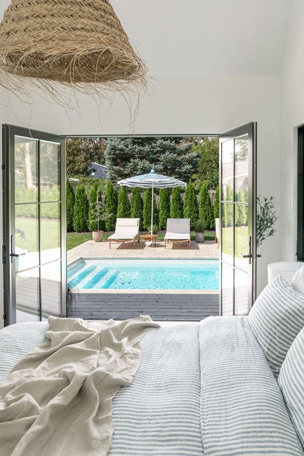 Two interior shots of a bedroom featuring a set of black E-Series Hinged Patio Doors that opens up to a deck and swimming pool outside.