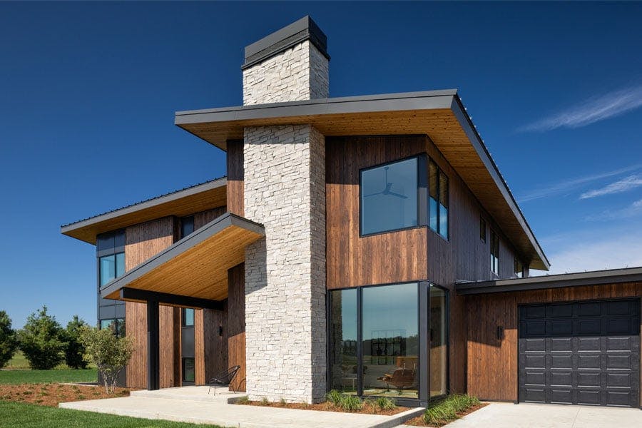 A contemporary home with vertical cedar cladding, long overhangs, a prominent stone chimney, and floor-to-ceiling black-framed windows.